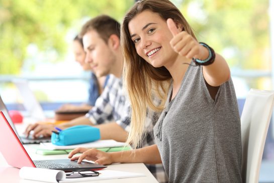 Iloinen opiskelija näyttää peukaloa kannettavan tietokoneen äärellä