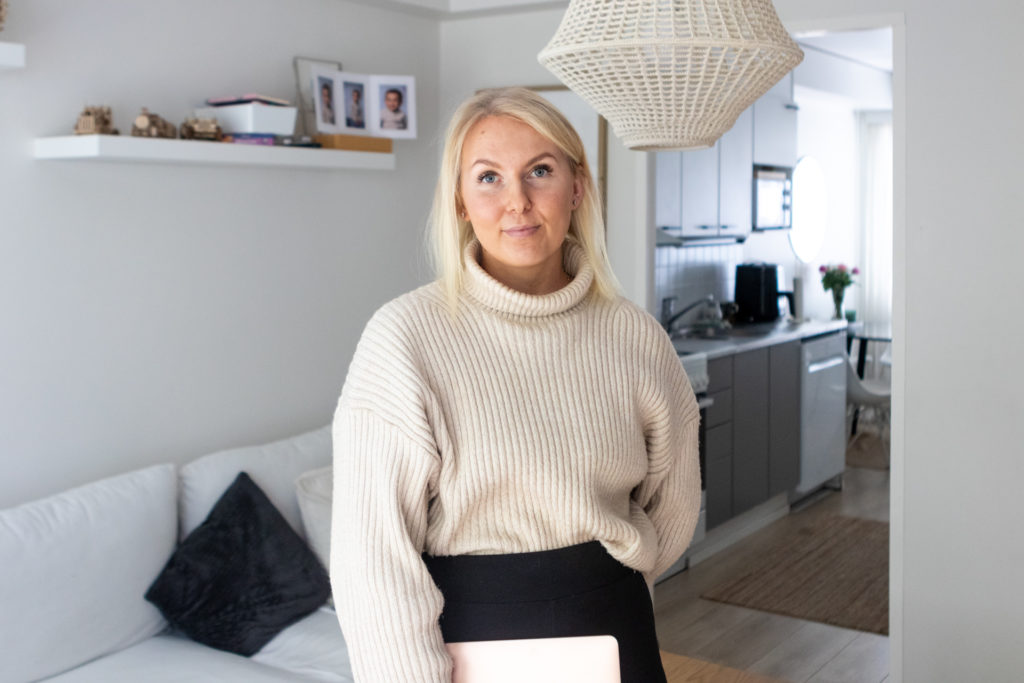 Linnea Lampi seisoo ja katsoo suoraan kameraan rivitalon olohuoneessa. Hänellä on yllään vaalea neule, mustat housut ja pitelee toisella kädellään Macbook Air -läppäriä. 