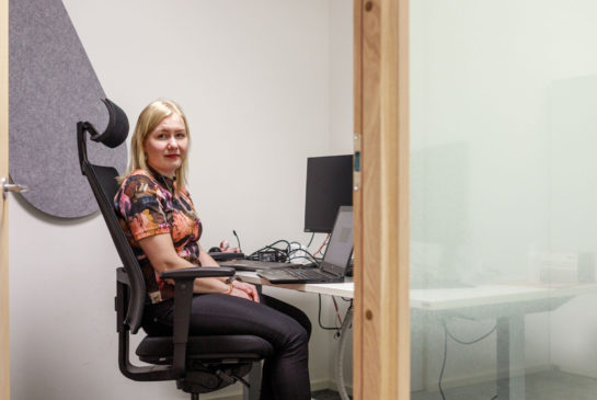 Annika Häkkinen istuu toimistolla työpöydän ja tietokoneen äärellä ja katsoo kameraan.