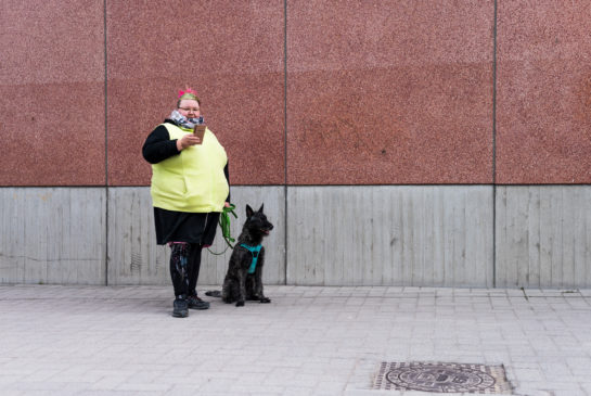 Punatukkainen nainen seisoo punatiiliseinää vasten keltaisissa huomiliiveisssä Itä-Helsingissä ja pitää mustaa koiraa hihnasta.