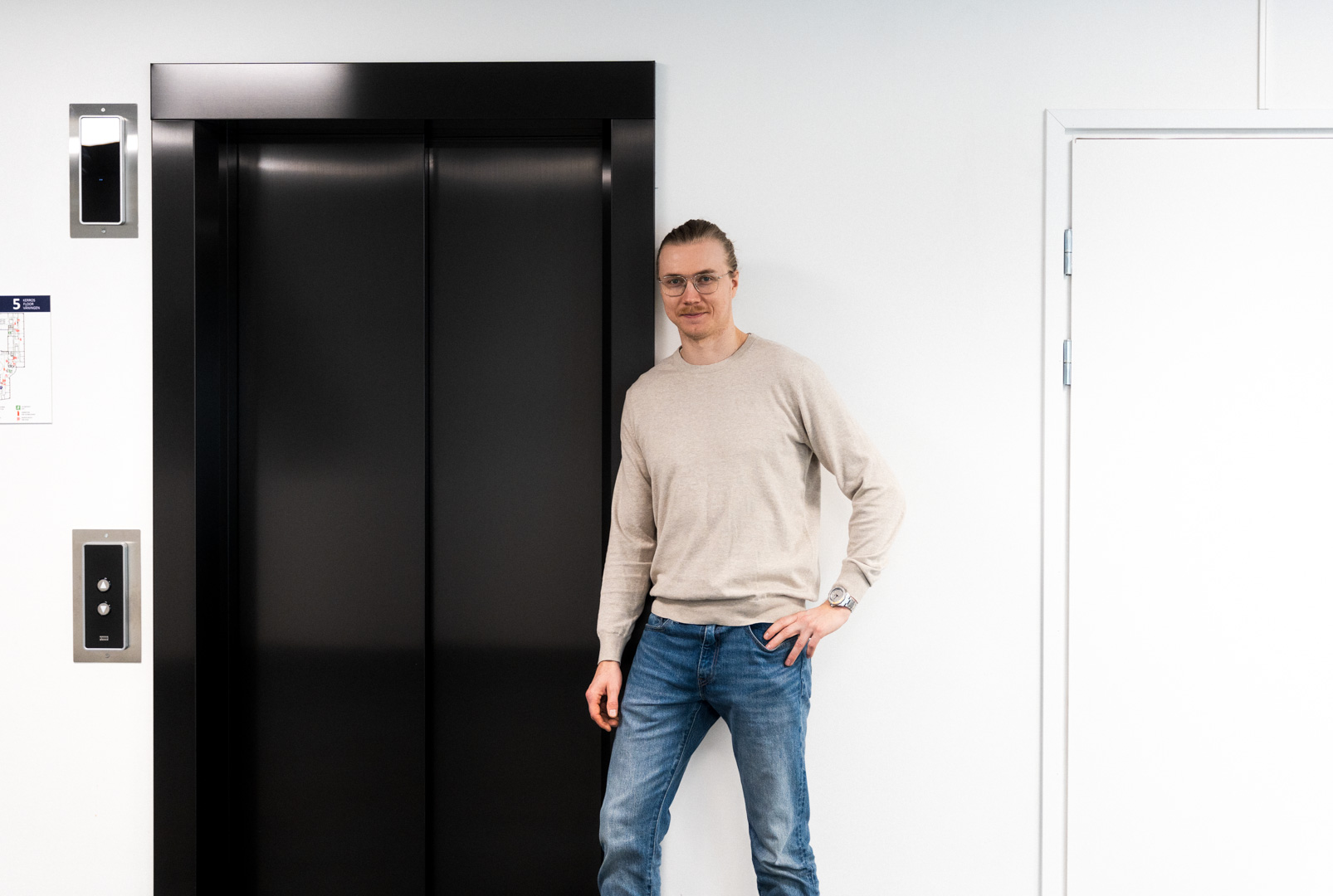 Tuomas Kaikkonen seisoo mustan hissinoven ja valkoisen seinä edessä.
