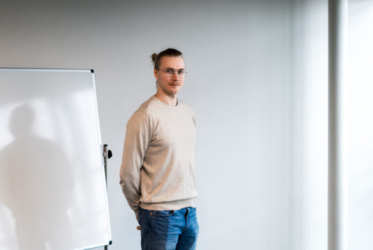 Tuomas Kaikkonen seisoo vaaleassa neuleessa ja sisinissä farkuissa valkoista toimistoseinää vasten.