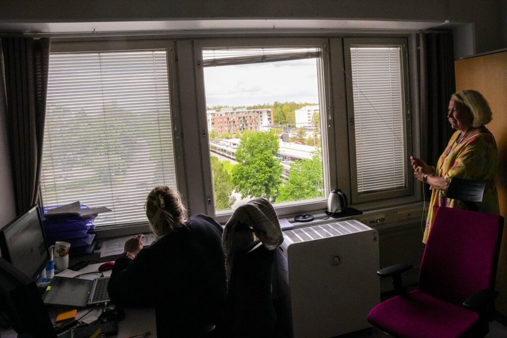 Laajakuva. Oikealla oleva Tolvanen avaa toimiston ikkunan sälekaihtimia. Ikkunasta näkyy ympäröivä ja vihertävä Martinlaakso. Kudd istuu työpisteellään kuvan vasemmassa laidassa.
